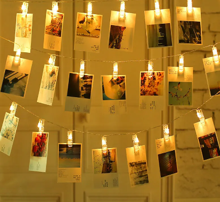 A festive and creative display of photographs and illustrations hung on a string of illuminated clip lights, spread across a wall as a part of room decor.