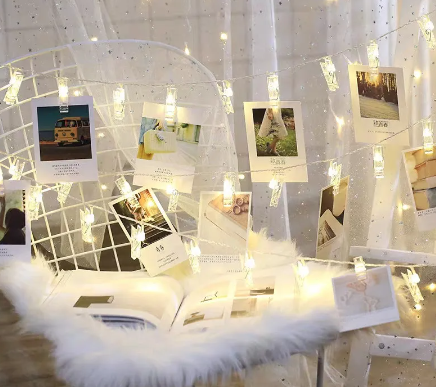 A string of clip lights hung against a white backdrop with several photographs clipped on, creating a decorative and personalized display of images.