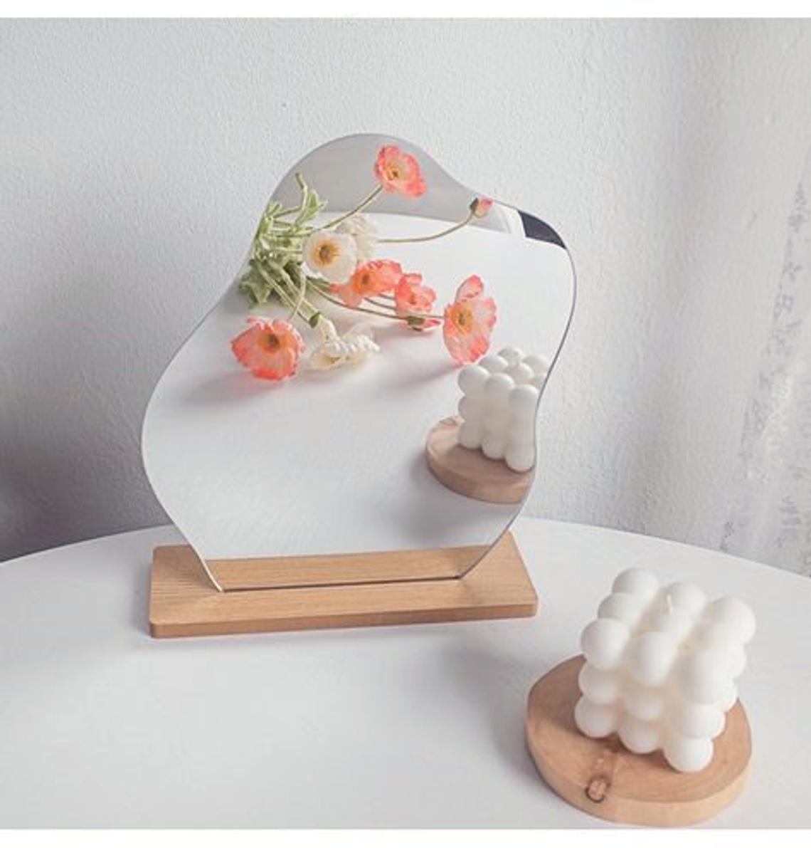 A wavy mirror with a curved top displayed on a wooden base, with a bouquet of orange flowers reflected in it, set on a white table next to a modern candle, fitting a cottagecore room decor.