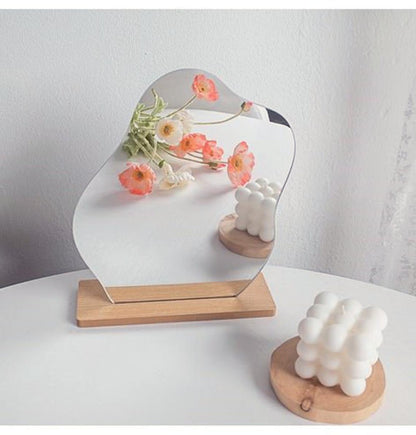 A wavy mirror with a curved top displayed on a wooden base, with a bouquet of orange flowers reflected in it, set on a white table next to a modern candle, fitting a cottagecore room decor.