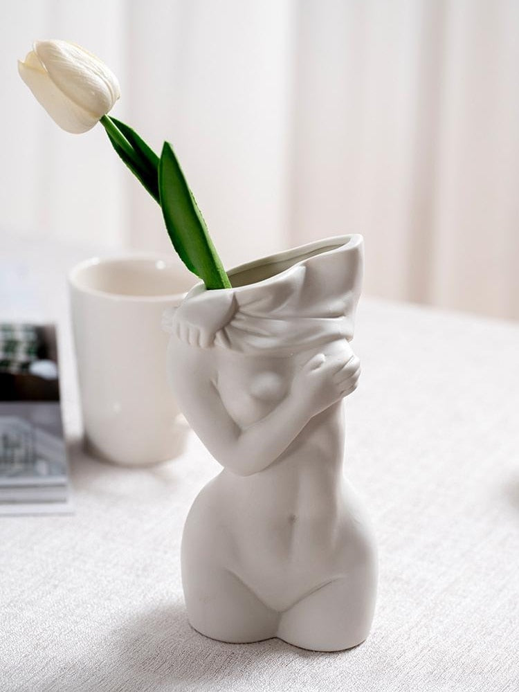 A white ceramic vase in the shape of a woman's torso with hands covering her face, holding a single tulip, placed on a table with a light curtain in the background.