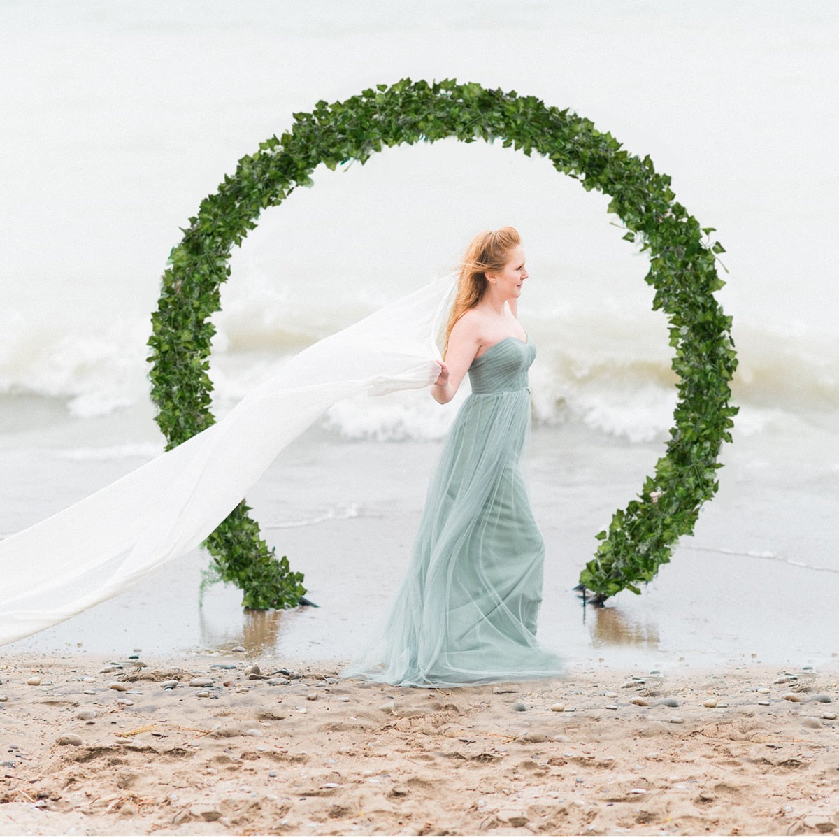 A bride on a beach with a large circular green ivy arch, creating a romantic and nature-inspired setting, ideal for a wedding or a cottagecore bedroom theme.