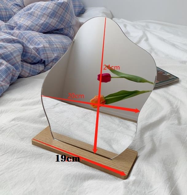 The wavy-edge mirror displayed in an upright position on a wooden stand, with dimensions marked as 24cm by 20cm, situated on a bed with a reflection of a tulip, suggesting a cozy and dreamy room decor.
