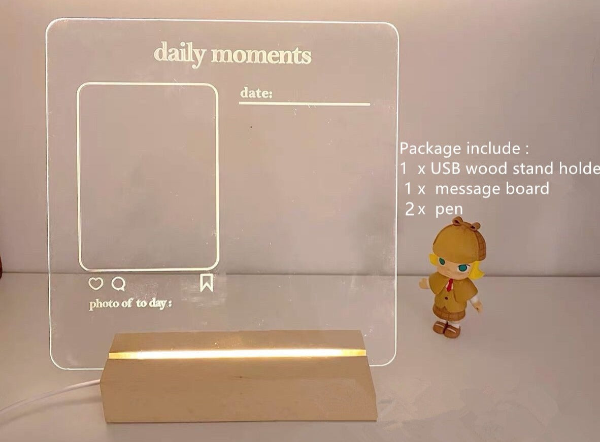 A simple and neat aesthetic LED memo board illuminated by soft light. The board is clear with a placeholder for a photo and the words "daily moments" and "date" printed on it. Accompanied by a small toy figure and the package content list on the side, it stands on a wooden USB-powered base.