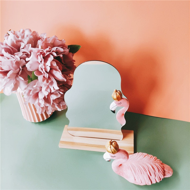 A cloud-shaped mirror standing on a wooden block with whimsical pink flamingo and gold ornament decorations beside a flower pot, reflecting a soft girl room decor aesthetic.