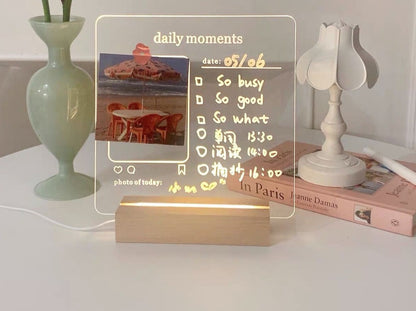 A personalized LED memo board with a photo and written notes detailing daily activities like "So busy" and "In Paris". The clear board is backlit by a soft glow, adding warmth to the scene which includes a classic lamp and a pastel book for a chic look.