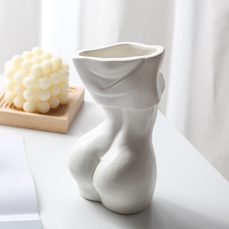 Another angle of the white ceramic vase, highlighting its curvaceous form and the draped cloth design at the top, set on a table with a decorative background.