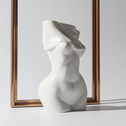 A white, matte, ceramic vase in the shape of a female torso with a draped cloth design around the upper part, positioned in front of a plain background with a copper frame to the side.