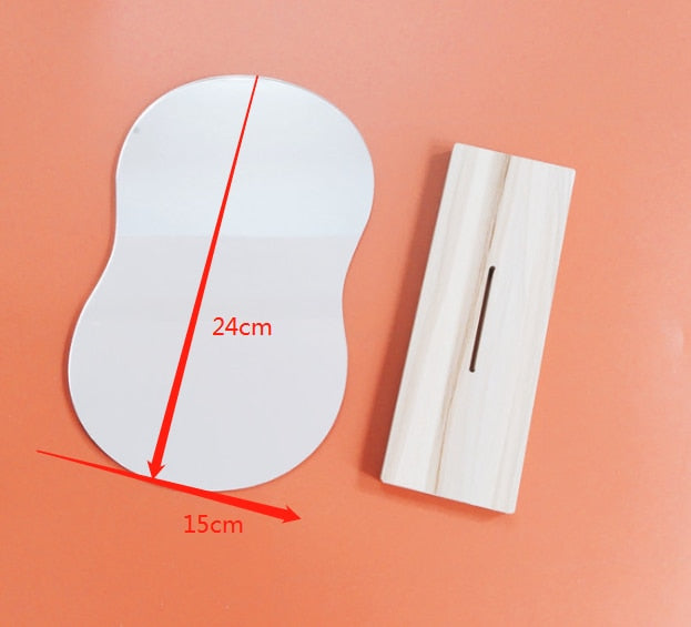 A model of a wavy-shaped mirror with dimensions of 24cm by 15cm laid flat next to a wooden stand for the mirror, on an orange background, embodying a minimalistic design.
