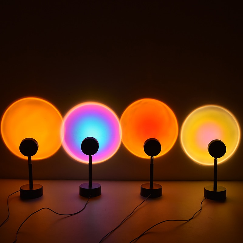 Four circular lamps on stands in a row projecting vibrant colored circles onto a wall; the colors are two shades of orange, a gradient of purple to blue, and yellow, resembling a sequence of eclipses.