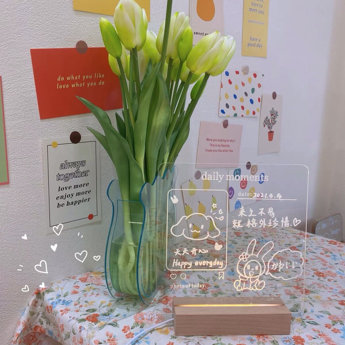 A vibrant LED memo board adorned with handwritten notes and drawings, highlighted by a warm backlight. The clear board, part of a colorful room decor, stands out amidst a bouquet of tulips and various decorative wall elements, creating an inviting and cheerful space.