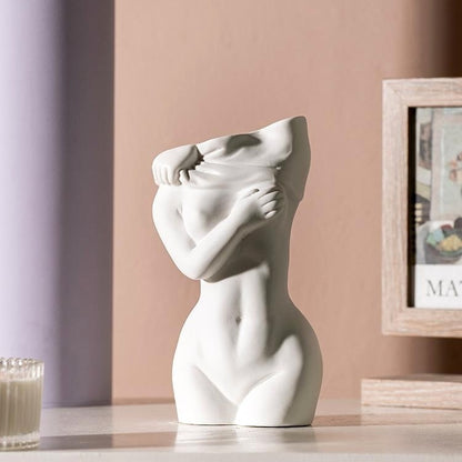 A white ceramic vase with a smooth finish, sculpted in the form of a woman's torso with hands covering her face, placed against a soft purple wall and next to a picture frame.