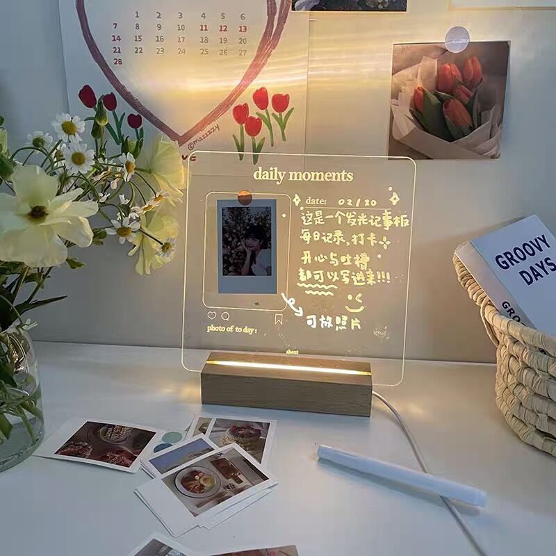 A charming LED memo board displaying a photo and handwritten notes under a soft light, creating a cozy atmosphere. The clear board features drawings and text celebrating "daily moments" and is accompanied by a stack of photos, enhancing the personal touch of the display.