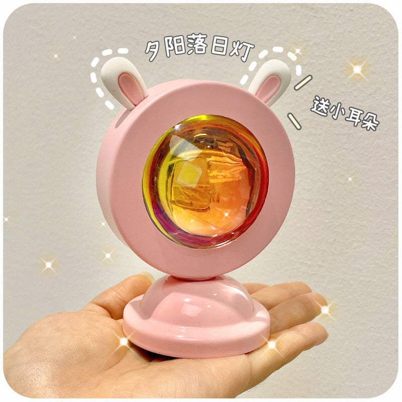 A hand holding a pink Sunset Light Projector with bunny ears design from the bedside lamps and dreamy room decor collection. The projector emits a warm, holographic glow, adding a cozy and cheerful room decor ambiance.