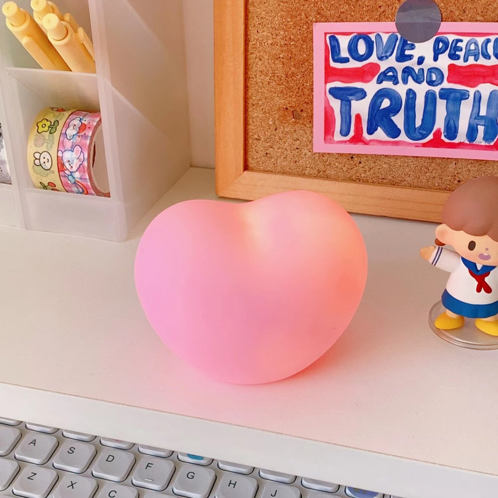 A vibrant pink heart-shaped lamp sitting on a desk next to a keyboard and a cute figurine, perfect for 'kawaii-room' and 'desk-accessories' collections, infusing a playful charm into a study or work area.