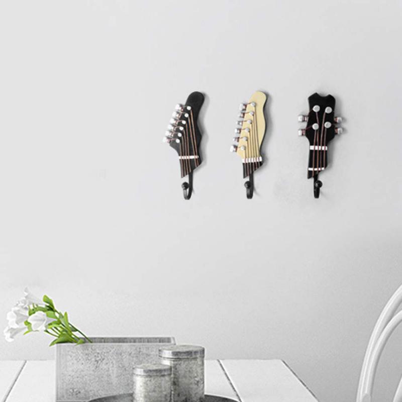 An image of a home interior wall featuring three wall hooks designed to mimic guitar headstocks. The left and right hooks are styled after black electric guitars, while the center one is cream-colored, resembling a classical guitar. A metal container with white flowers is placed on a white table in the foreground, providing a cozy domestic ambiance.