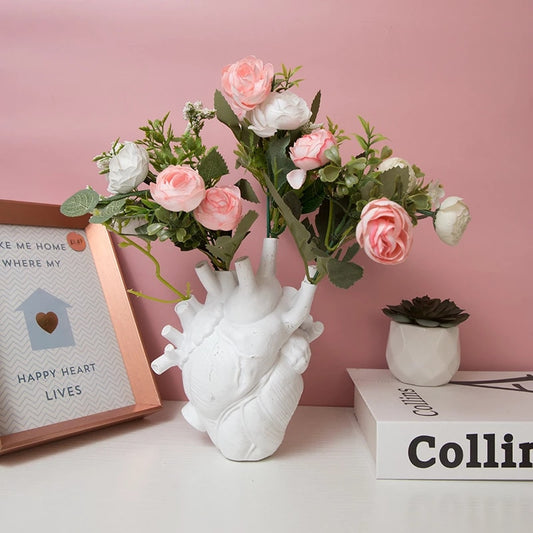 A white heart-shaped vase filled with artificial pink roses is placed on a desk, illustrating the vase's potential as an endearing home decor item.