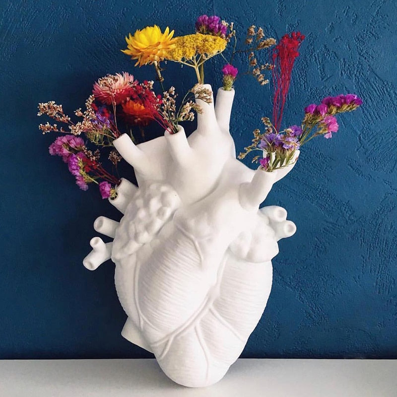 A white ceramic heart-shaped vase serves as a planter for colorful flowers including yellow, red, and pink blooms against a deep blue wall.