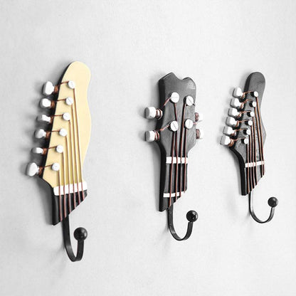 A close-up view of three decorative wall hooks on a light gray wall, each designed in the style of guitar headstocks with detailed tuning pegs and strings. The first hook on the left is designed like a classical guitar in cream color, the middle is a darker, modern electric guitar headstock, and the third resembles a dark acoustic guitar headstock, all with metallic tuning pegs.
