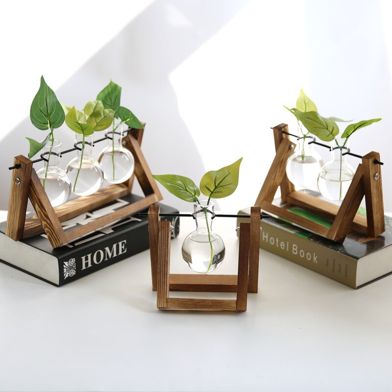 An array of wooden terrarium stands featuring clear bulb vases with green plants. Each stand has a triangular frame and holds three vases, set against a bright background with books titled 'HOME' and 'Hotel Book,' suggesting a cozy and educational atmosphere, suitable for desk accessories or a cottagecore room decor.