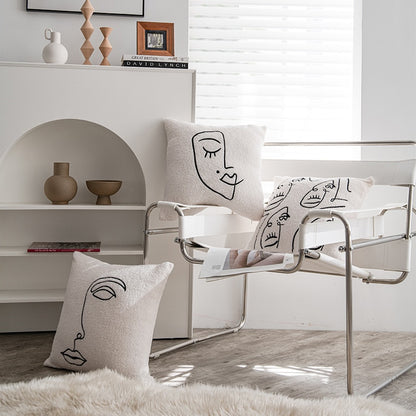 A well-lit, serene room featuring three pillows with abstract line drawings of faces, placed on a modern chair and a fluffy rug. The decor presents a minimalist aesthetic, complementing the 'Art Hoe' style pillowcases. The background showcases a white shelf with neutral-toned vases, books including one titled 'Great Britain David Lynch,' contributing to the room's 'Boho', 'Coquette', 'Cottagecore', 'Danish Pastel', 'Dorm Room', 'Dreamy', and 'Relaxing' decor themes.