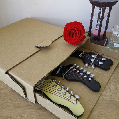 An open cardboard box reveals three guitar headstock-shaped wall hooks nestled inside. Two black electric guitar designs flank a cream-colored classical guitar design in the center. A single bright red rose rests on the edge of the box, adding a touch of color to the scene, which includes a wooden surface and a blurred background hinting at a cozy interior space.
