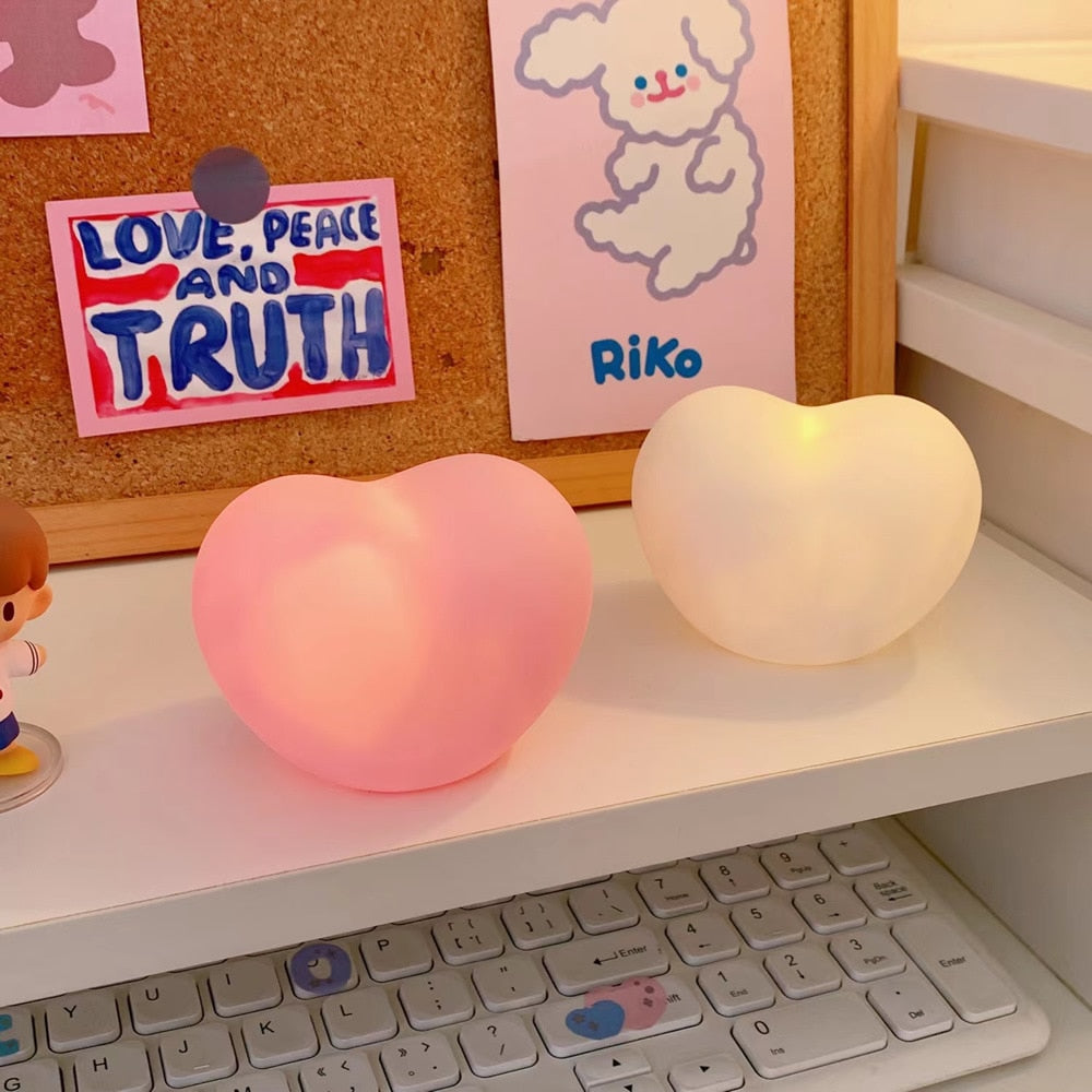 Two heart-shaped lamps, one pink and one white, glow softly on a desk setup, complementing the 'desk-lamps' and 'feminine-room-decor' collections, with playful stationery and a keyboard in the background, suggesting a warm, inviting study space.