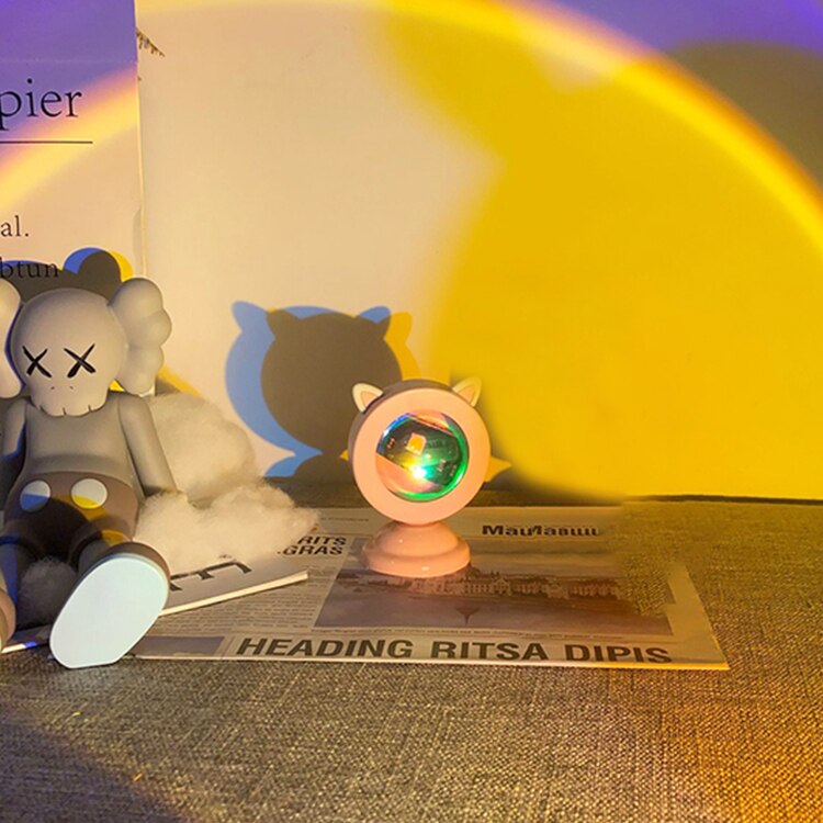 A Sunset Light Projector against a background of gradient yellow to violet hues, next to a plush toy, creating a soft ambiance suitable for a soft girl room or a y2k room decor theme. The projector's light mimics the golden hour, perfect for photography and creating a cozy atmosphere.