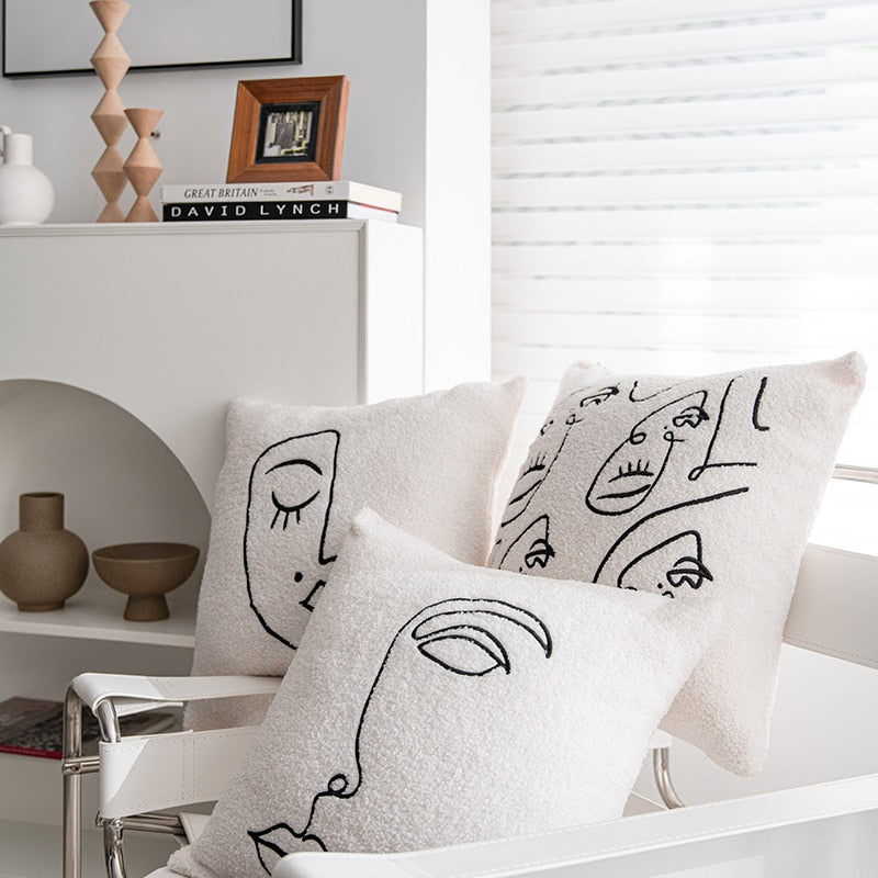 A contemporary, airy room featuring two artfully designed pillowcases on a sleek metal chair, each with a unique black line drawing of a face, epitomizing the 'Art Hoe' aesthetic. The background includes a white shelf adorned with vases and a book stack topped with a 'Great Britain David Lynch' book, harmonizing with the 'Boho', 'Coquette', 'Cottagecore', 'Danish Pastel', 'Dorm Room', 'Dreamy', 'Feminine', and 'Relaxing Room' decor themes.
