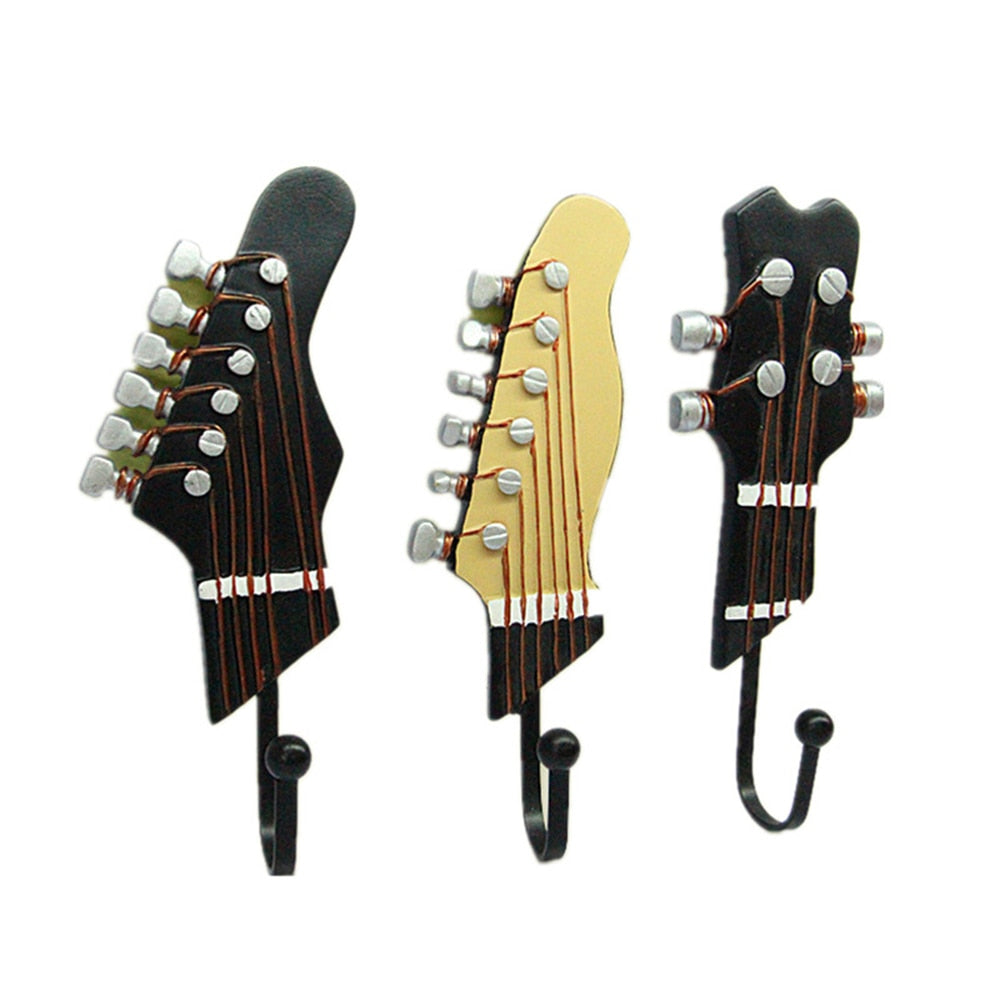 Three decorative wall hooks isolated on a white background, crafted to resemble the headstocks of guitars with realistic details such as tuning pegs and strings. From left to right: a black electric guitar headstock design, a cream-colored classical guitar headstock, and another black electric variant, all with six pegs indicative of six-string instruments.