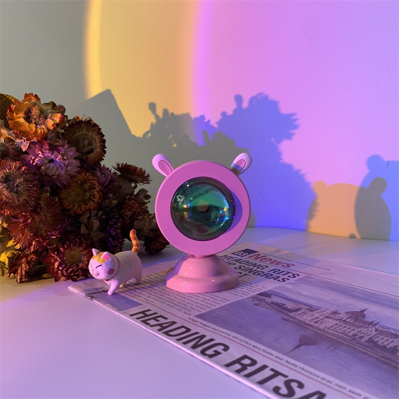 Aesthetic sunset light projector with cat ears casting a soft shadow against a gradient wall, placed on a newspaper, complementing a boho-room decor and bedside lamps collection with a dried flower bouquet adding a cottagecore touch.