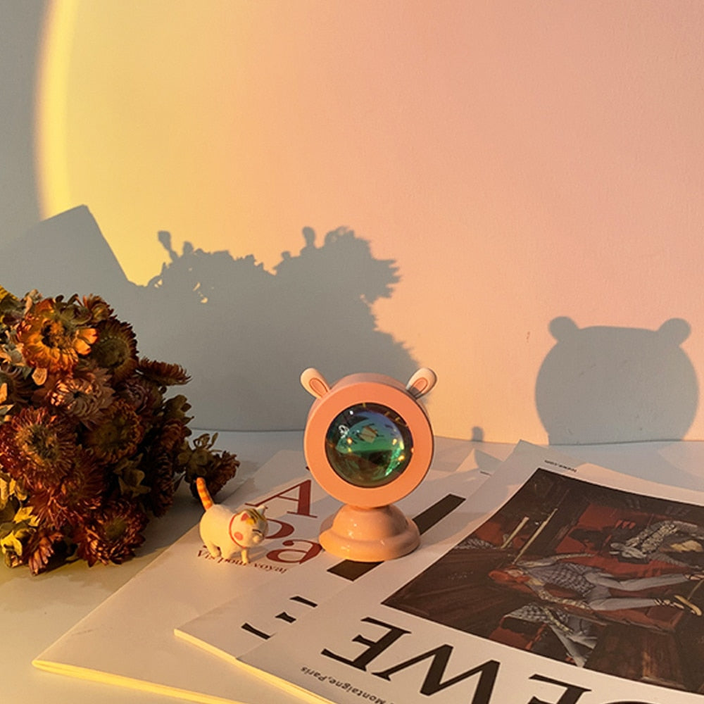 A peach-colored Sunset Light Projector with cat ears design on a white surface, casting a warm sunset glow against a plain wall. The scene is accessorized with dried flowers and a magazine, creating a cozy art hoe and cottagecore room decor ambiance
