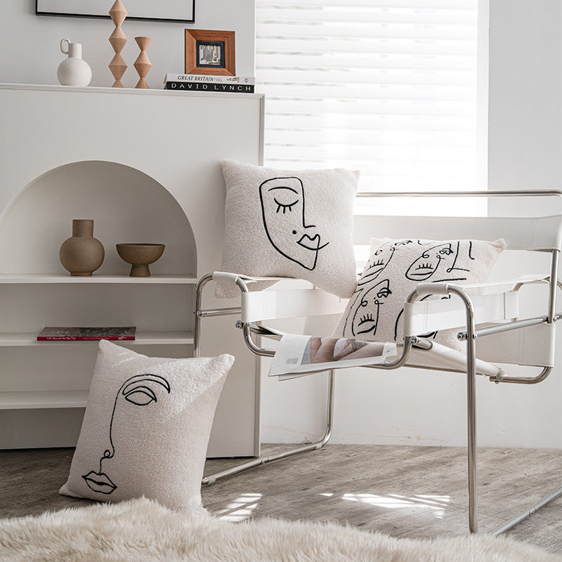 A stylish room featuring a modern metal chair on which rest two pillowcases with abstract face drawings, and a third pillowcase on a fluffy white rug. In the background, a white shelf holds simple decor items and a book with 'David Lynch' on the spine. This setting evokes a serene and artistic vibe, fitting for 'Art Hoe' room decor and aligning with themes such as 'Boho', 'Coquette', 'Cottagecore', 'Danish Pastel', 'Dorm Room', 'Dreamy', 'Feminine', and 'Relaxing Room' decor.