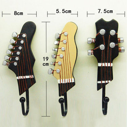 Three guitar headstock-shaped wall hooks displayed against a cream background, each with specific dimensions labeled. The leftmost, a black electric guitar headstock, measures 8 cm wide, the center, a cream-colored classical guitar headstock, measures 5.5 cm wide, and the rightmost, another black electric guitar headstock, is 7.5 cm wide. All are 19 cm in height, showcasing their size and design for potential buyers.