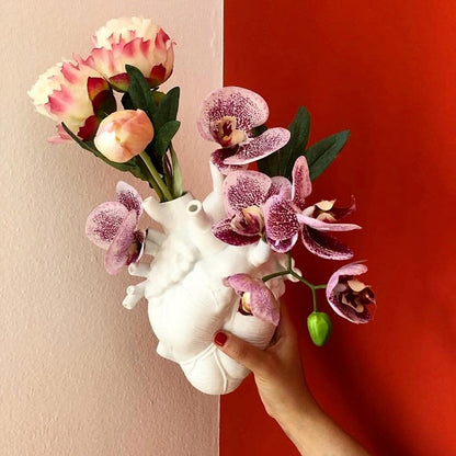 A person holds a white ceramic vase modeled in the shape of a human heart, filled with a vibrant arrangement of pink tulips and purple orchids against a red background.