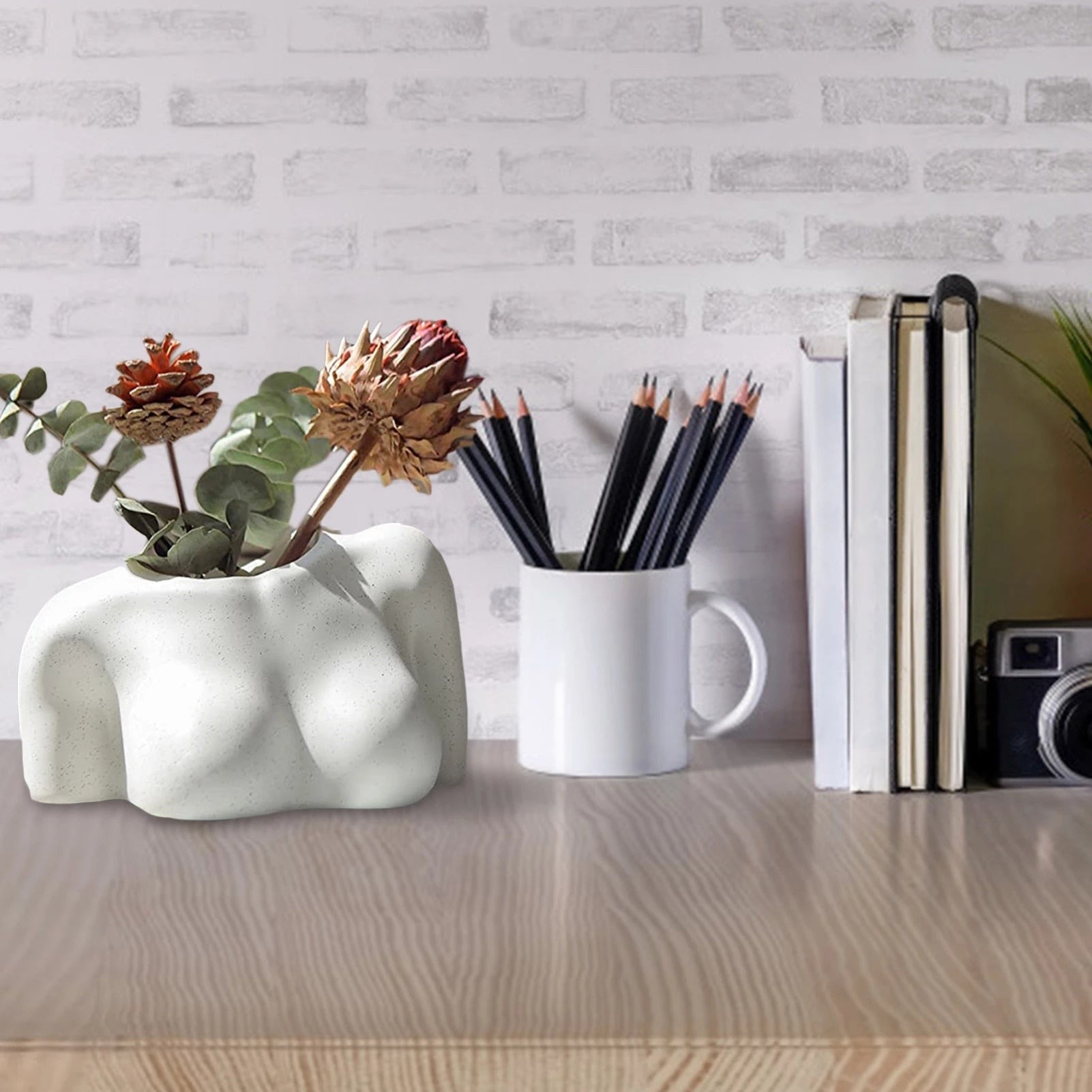The speckled white vase placed on a desk next to a cup holding pencils, books, and a camera, blending the vase into everyday life as a functional decor item.