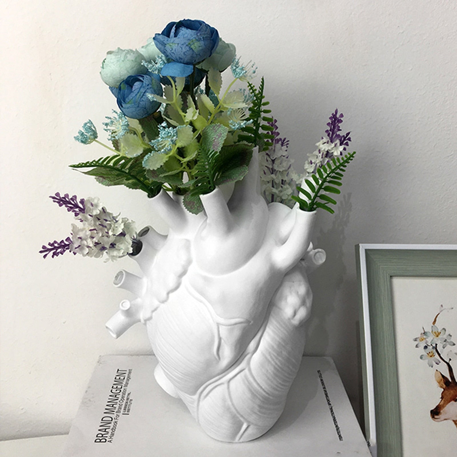The heart-shaped vase is styled with light blue flowers and green foliage, displayed on a shelf next to a picture frame, showcasing its decorative and functional design.
