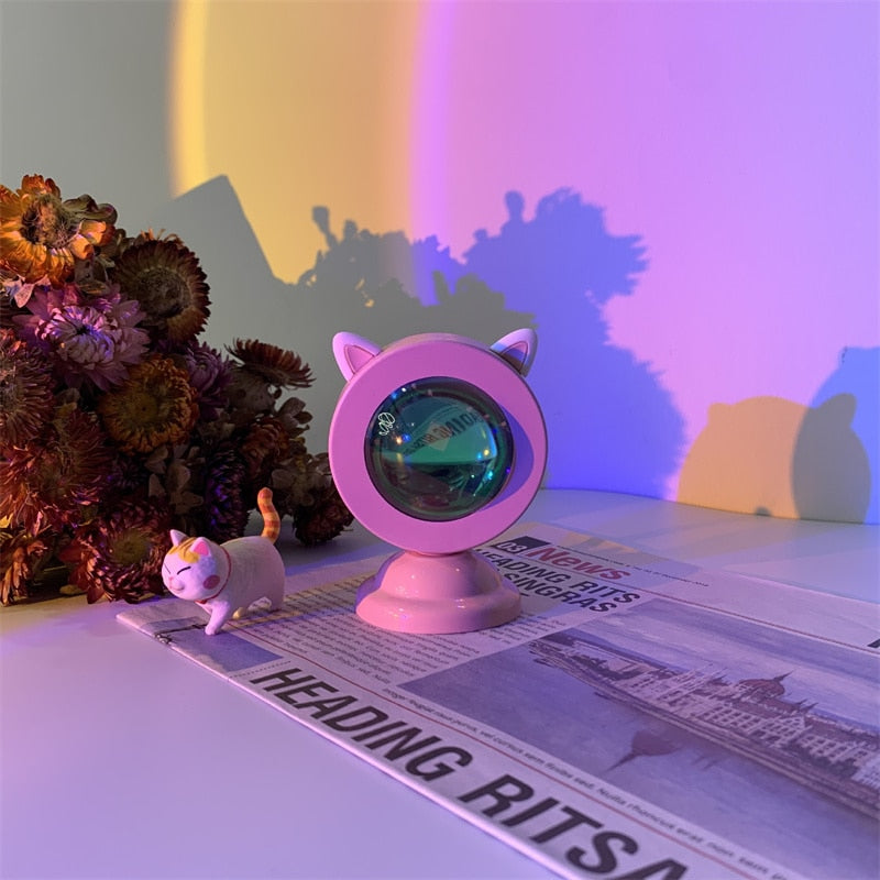 A Sunset Light Projector in a pink hue with cat ears design, illuminating a room with a blend of purple and orange light. Perfect for adding a touch of kawaii room or boho room decor, the projector sits beside a quirky figurine and atop a newspaper, suggesting a playful yet serene setting.