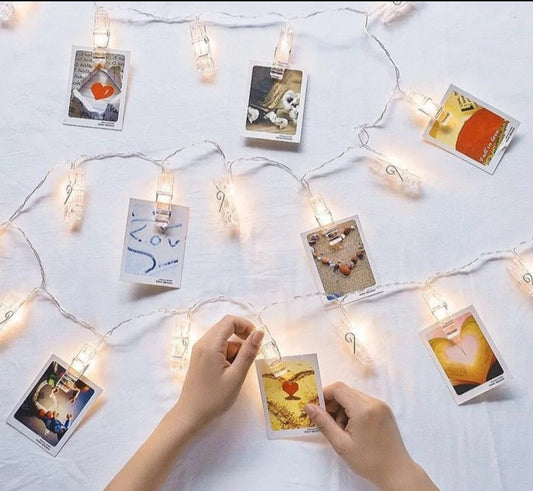 An interactive scene where a person's hands are seen placing a photograph onto a string of clip lights spread across a white surface, with other photos already attached to the glowing clips.