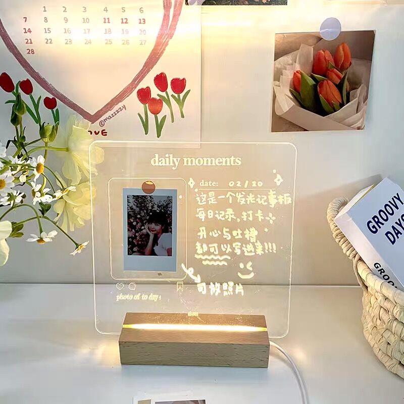 A warmly lit aesthetic LED memo board with a photo of a smiling person clipped to it. The clear board is marked with handwritten notes and drawings, with the caption "daily moments" and a "photo of the day" section. Decorated with a faux flower arrangement and a 'GROOVY DAY' book, the scene exudes a cozy ambiance.