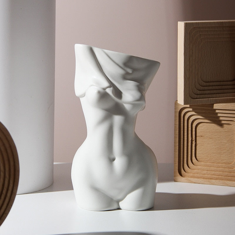 A matte white ceramic vase with an artistic design, sculpted to resemble the female form with a flowing fabric effect around the upper portion, displayed against a minimalistic background with soft shadows.
