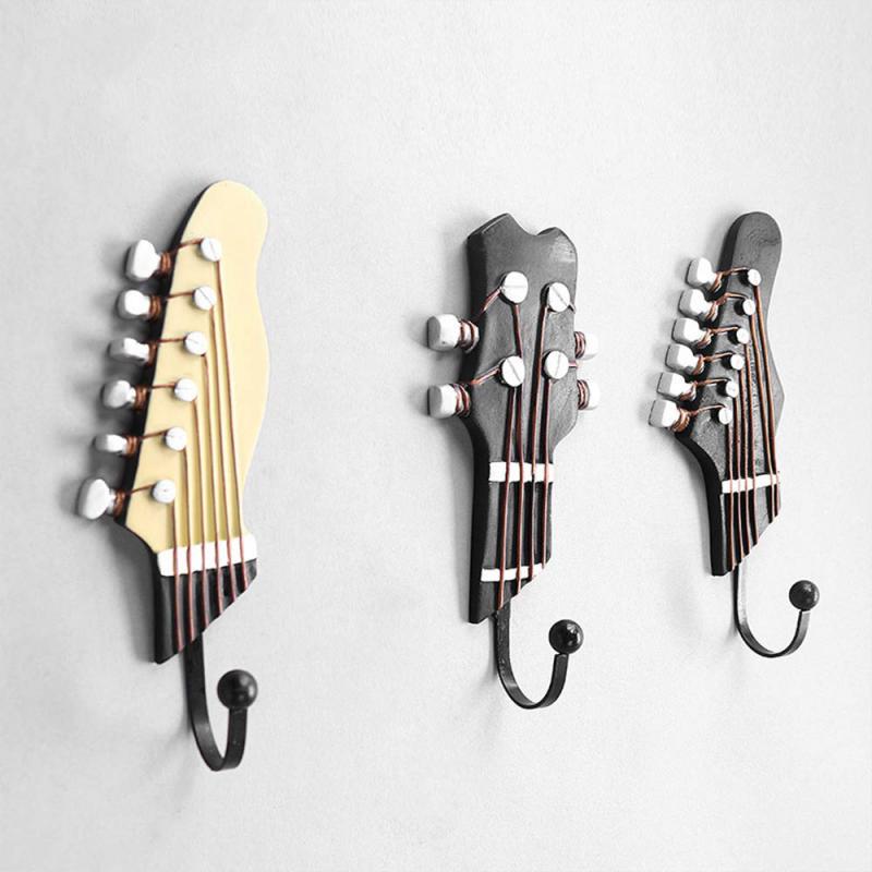 Three wall hooks designed to look like the headstocks of stringed instruments, with tuning pegs and strings, mounted on a light gray wall. The leftmost hook is cream-colored, resembling a classic guitar headstock, while the two on the right are darker, imitating a more modern design, each with six pegs for tuning.